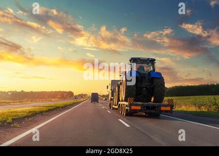 POV schweren Flurförderzeug Sattelauflieger Flachbett Plattform Transport zwei große moderne Landwirtschaft Traktor Maschine auf gemeinsame Autobahn Straße bei Sonnenuntergang Sonnenaufgang Himmel Stockfoto