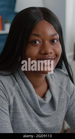 Porträt einer afroamerikanischen Studentin, die am Schreibtisch im Wohnzimmer sitzt und während der Heimsuchung lächelnd in die Kamera blickt. Schwarzer Teenager, der Informationen zur Schule durchsucht und von zu Hause aus arbeitet Stockfoto