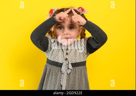 Kleines Mädchen in einem grauen Kleid auf gelbem Hintergrund. Hände über den Kopf symbolisieren den Kauf eines neuen Hauses. Stockfoto