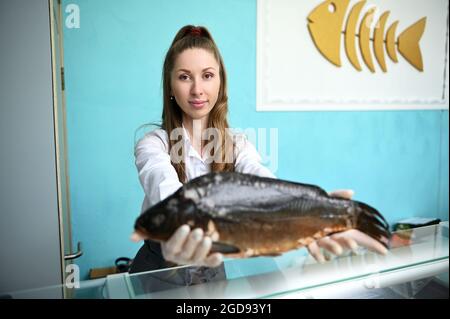 Eine hübsche weibliche Fischmongerin hinter der Theke, die einen riesigen Fisch in ihren ausgestreckten Händen hält. Fischgeschäft im Fischgeschäft Stockfoto