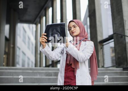 Weibliche Ärztin aus dem Nahen Osten, die Hijab und weißen Mantel mit Stethoskop trägt, draußen auf dem Hintergrund der Treppe eines modernen Krankenhauses steht und das Röntgenbild betrachtet Stockfoto