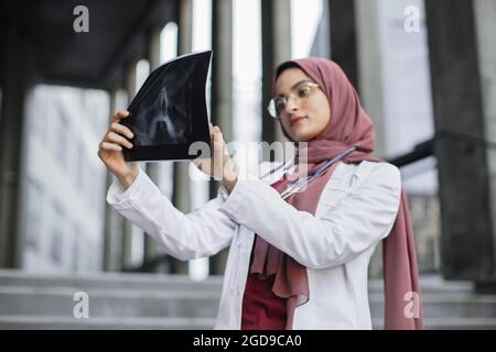 Röntgenbild, Roentgenbild. Seriöse, nachdenkliche muslimische Chirurgen, die sich die Röntgenradiographie des Schädels des Patienten anschaut. Weibliche arabische Ärztin überprüft Röntgenscan im Freien. Selektiver Fokus auf Röntgenscan Stockfoto