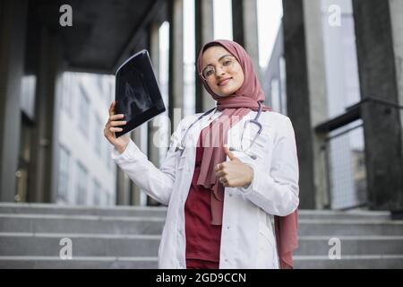 Attraktive, glückliche muslimische Ärztin in Hijab, untersucht eine Röntgenaufnahme des Schädels des Patienten und lächelt mit dem Daumen nach oben auf die Kamera. Arabische Ärztin, die im Freien mit Röntgenscan steht Stockfoto