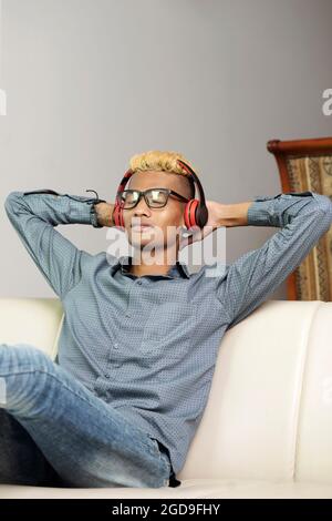 Hübscher junger Mann mit Brille, der sich auf dem bequemen Sofa zurücklehnt, die Augen schließt und in Kopfhörern Musik hört Stockfoto