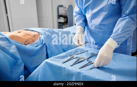 Chirurg in sterilen Handschuhen, die medizinische Instrumente vorbereiten. Patient mit Flecken auf der Haut, die auf dem Bett liegen, während der Arzt Scheren, Pinzetten und Skalpelle vorbereitet. Konzept der Vorbereitung der plastischen Chirurgie. Stockfoto