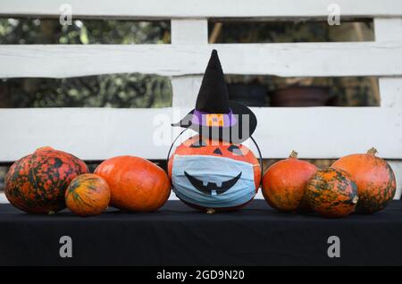 Halloween-Dekorationen in der neuen Realität der COVID-19 Pandemie. Reihe von Kürbissen, von denen einer Hexenhut und medizinische Maske mit bemalten gruseligen Lächeln hat. St Stockfoto