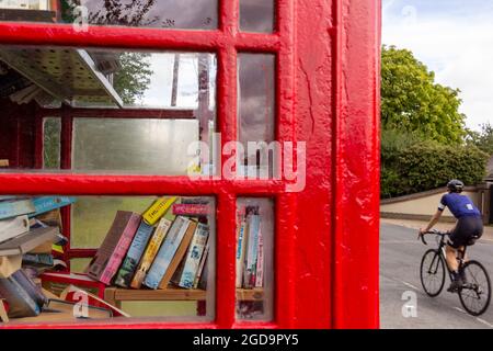 Beim Blick durch das Fenster einer stillgelegten öffentlichen Telefonbox verwendete man jetzt eine Bibliothek, wobei der Radfahrer im Hintergrund auf der Straße vorbeikam Stockfoto