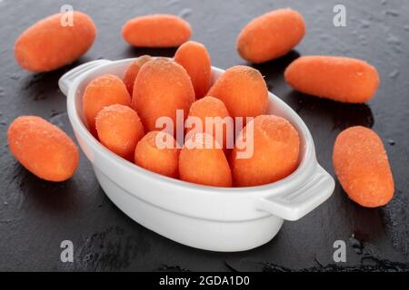 Mehrere leuchtend orange saftige, natürliche Mini-Karotten in einer weißen Keramikschale, Nahaufnahme, auf einem Schiefertafel. Stockfoto