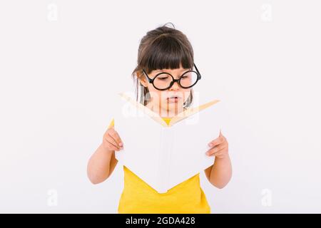 Das kleine Mädchen war überrascht und trug ein gelbes T-Shirt und eine runde schwarze Brille, die ein offenes Buch in den Händen hielt und las, auf weißem Hintergrund Stockfoto