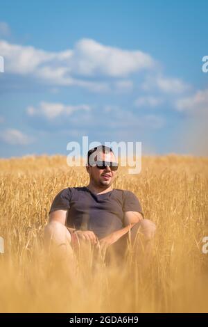 Ein junger Mann mit Sonnenbrille sitzt an einem sonnigen Sommertag mitten auf einem Triticale-Feld. Konzept der Landwirtschaft und Wirtschaft. Vertikales Foto Stockfoto