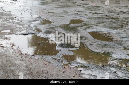 Ein mit Wasser gefülltes Loch in einer asphaltierten Straße. Ein regnerischer Tag in einer Großstadt fahren Autos auf der alten Straße. Schlechte Straße mit Asphalt in Gruben und Schlaglöchern tha Stockfoto