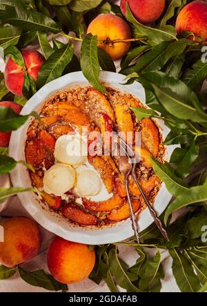 Hausgemachter Pfirsich-Crisp mit Eis, umgeben von frischen rohen Pfirsichen Stockfoto