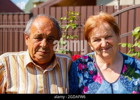 Porträt eines älteren Paares, das am Sommertag lächelt. Glückliches älteres Paar von Mann und Frau sitzen auf Blumen Garten Hintergrund. 60er oder 70er Jahre Mann und woma Stockfoto