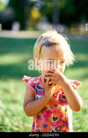 Kleines Mädchen isst einen Pfirsich auf einem grünen Rasen. Hochformat Stockfoto