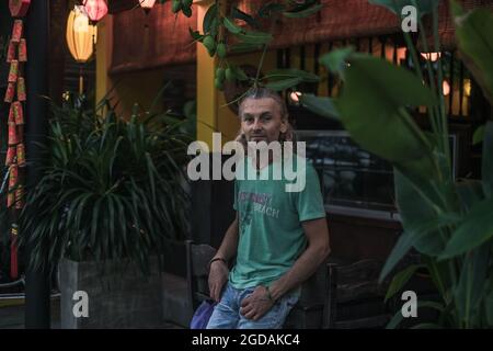 Älterer Mann in T-Shirt, Jeans in der Nähe des asiatischen Restaurants. Umgeben von tropischen Pflanzen. Lange graue Haare. Chinesische Laternen. Abendportrait. Stockfoto