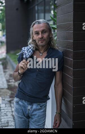 Älterer Mann in T-Shirt, Jeans, die in der asiatischen Stadt auf der Straße bleiben und seine Haare halten. Lange graue Haare. Modernes Haus. Hochwertige Fotos Stockfoto