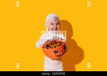 Kind im Mumienkostüm mit Pralinenkorb vor gelbem Hintergrund. Halloween Trick or Treat Stockfoto