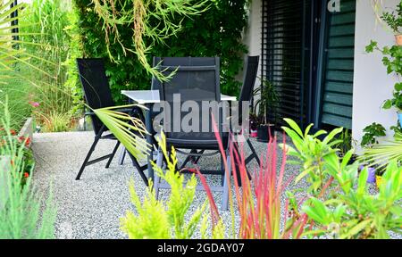 Wunderschöne Außenterrasse mit Steinteppichboden, wachsenden grünen Blumen und Pflanzen und Tisch mit Stühlen. Haus Terrasse mit Pflanzen überwuchert Stockfoto