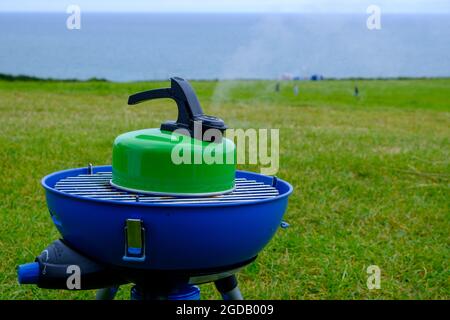 Blauer Campingaz Campingkocher mit einem grünen Wasserkocher mit Dampf aus dem Auslauf und das Meer im Hintergrund.2021 Stockfoto