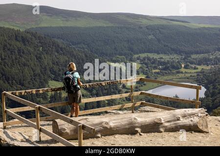 Wanderfrau mit Blick auf die Glendalough Seen von einem Aussichtspunkt aus mit Graffiti am Zaun, auf der Spinc Walking Route, County Wicklow, Irland Stockfoto