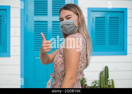 Geimpfte glückliche kaukasische junge Frau mit blonden Haaren, die eine Einweg-Gesichtsmaske trägt und den Daumen zeigt. Covid-19- oder Coronavirus-Impfstoff. Impfung Stockfoto
