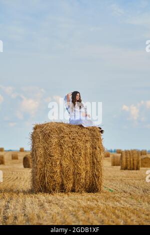 Verträumte junge Frau sitzt auf dem Strohstapel auf dem Feld Stockfoto