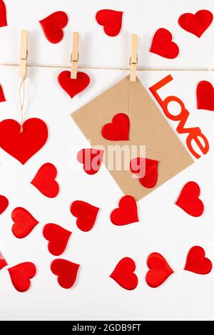 Hölzerne Wäscheklammern hängen an einem Seil auf weißem Hintergrund und halten ein Herz und einen Umschlag. Viele verstreute Herzen und das Wort Liebe. Selektiver Fokus Stockfoto