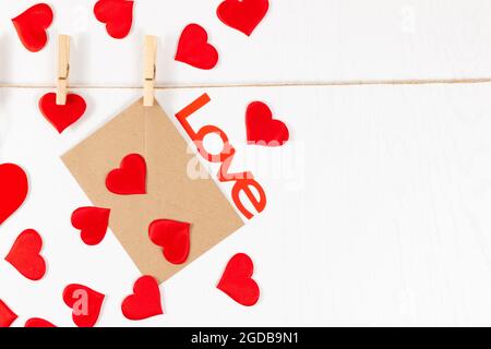 Hölzerne Wäscheklammern hängen an einem Seil auf weißem Hintergrund und halten ein Herz und einen Umschlag. Viele verstreute Herzen und das Wort Liebe. Selektiver Fokus Stockfoto