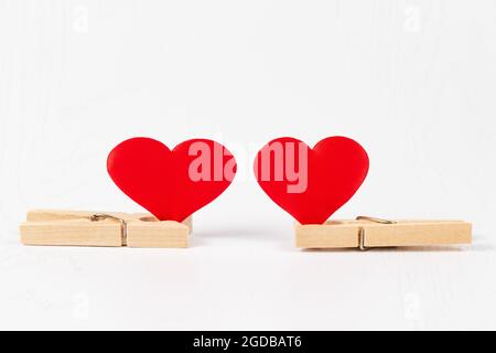 Zwei hölzerne Wäscheklammern halten zwei rote Herzen auf einem weißen Holztisch Hintergrund Nahaufnahme. Selektiver Fokus Stockfoto