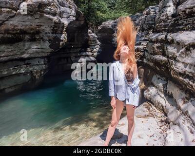 Das langhaarige Mädchen wirft ihren Kopf zurück, während es am türkisfarbenen Wasser an einem felsigen Ufer zwischen den Schluchten steht Stockfoto