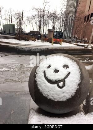 Smiley-Gesicht mit Zunge, die im Schnee auf einer großen Zementkugel in Lower Manhattan, New York City, ragt. Stadtpark im Hintergrund. Stockfoto