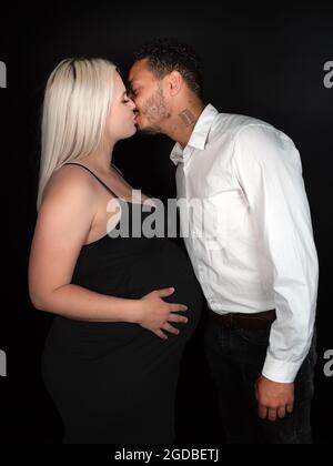 Profilansicht eines stilvollen und glücklichen multiethnischen Paares, das ein Baby erwartet und sich liebevoll küsst Stockfoto