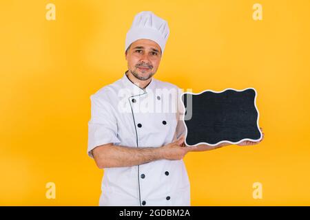 Chefkoch trägt weiße Küchenmütze und Jacke mit schwarzer Kreidetafel auf gelbem Hintergrund Stockfoto