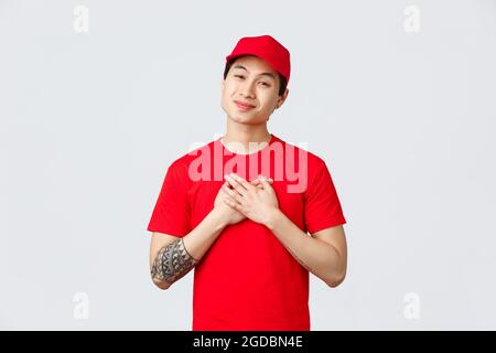 Express-Versand- und Logistikkonzept. Berührte glücklich asiatischen Lieferung Kerl in roten Kappe und T-Shirt, halten Sie die Hände auf Herz, seufzend und starrte Stockfoto