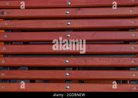 Die Rückseite einer Holzbank, braun lackiert, bestehend aus horizontal befestigten schmalen Lamellen, in der Mitte mit Schrauben am Metallsockel befestigt Stockfoto