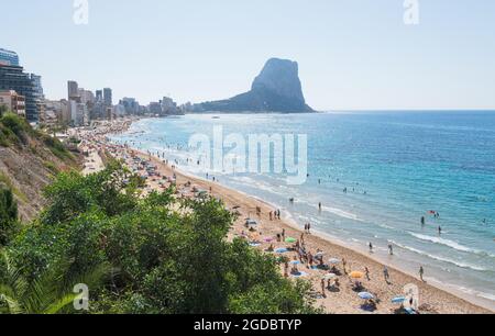 Calpe, Spanien - 19. Juli 2021: Sandstrand mit vielen Touristen für Freizeit, Schwimmen und Sonnenbaden entlang der Stadt Calpe mit Blick auf den Felsen formati Stockfoto