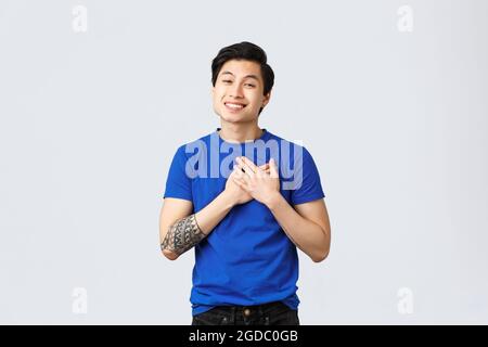 Berührt und dankbar schöner männlicher Asian in blauem T-Shirt mit Tattoos, berührendes Herz und Lächeln, schätzen Hilfe, erfreut oder erfreut sein Stockfoto