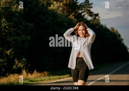 Eine rothaarige Frau in einem weißen Hemd geht an einem sonnigen Sommertag entlang der Straße durch den Wald. Ein Ausflug außerhalb der Stadt. Stockfoto