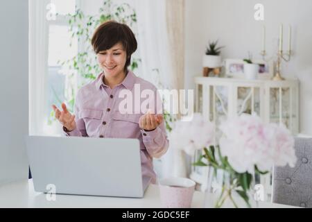 Geschäftsfrau, die zu Hause arbeitet, einen Laptop benutzt, gestikiert, an einem Online-Meeting teilnimmt. Gemütlicher Arbeitsplatz mit frischen Pfingstrosen-Blüten auf Weiß Stockfoto