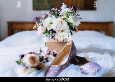 Brautstrauß mit frischem Eukalyptus, Pfingstrosen und Onustomblumen elegant mit Hochzeitsschuhen und floralem Haardekor auf dem Bett im Fine Art Stil. Selektiv Stockfoto