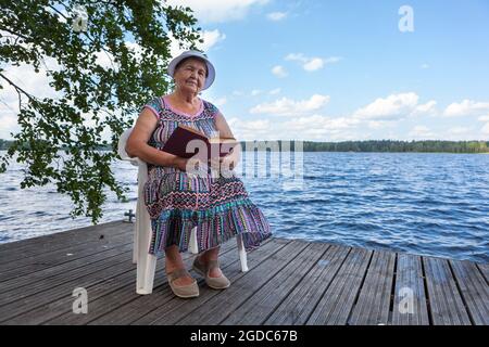 Eine alte Dame, die ein Buch liest, sitzt am Plastikstuhl am Waldseeufer und schaut auf die Kamera Stockfoto
