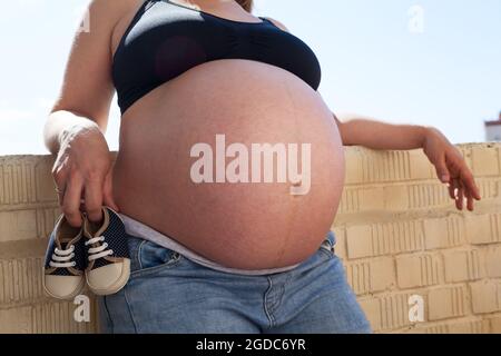 Kleine Schuhe für das ungeborene Baby im Bauch einer schwanger kaukasischen Frau Stockfoto