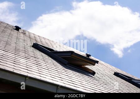 Ein Porträt eines offenen Dachfensters zur Belüftung in einem Schiefer-Dach neben einem anderen an einem sonnigen Tag mit einem blauen Himmel mit weißen Wolken. Sie können al Stockfoto
