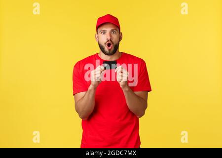 Kontaktloses Bezahlen, Online-Lieferung, Internet-Shopping-Konzept. Aufgeregt und erstaunt junger männlicher Mitarbeiter in Firmenuniform, rotem T-Shirt und Mütze Stockfoto