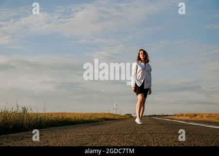 Eine rothaarige Frau in einem weißen Hemd geht an einem sonnigen Sommertag entlang der Straße zwischen den Feldern. Ein Ausflug außerhalb der Stadt Stockfoto