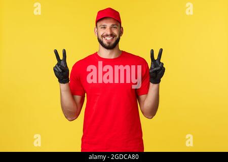 Express-Postversand, Paketübertragung und Logistikkonzept. Freundlicher netter Kurier in roter Uniform, zeigt Friedenszeichen in Schutzhandschuhen, lächelnd Stockfoto