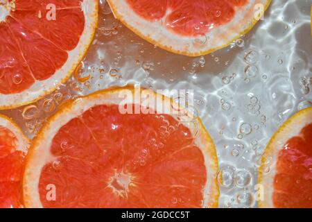 Nahaufnahme der roten Grapefruitscheiben im Limonadenhintergrund. Textur des kühlenden süßen Sommergetränks mit Makroblasen an der Glaswand. Flach Stockfoto
