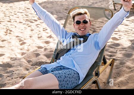 Der glückliche junge Unternehmer downshifter freut sich, auf einer Sonnenliege am Strand zu liegen, bekleidet mit Hemd, Krawatte und Unterhose, und hebt die Hände hoch Stockfoto