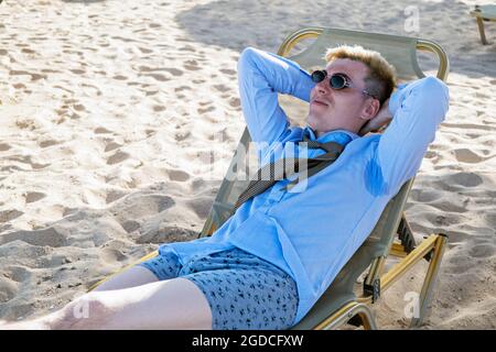 Der junge Mann liegt auf einer Sonnenliege mitten am Strand in einem Hemd mit Schweißflecken auf seinen Achselhöhlen, Krawatte und Shorts und wirft seine Hände hinter seine Stockfoto