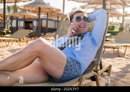 Der junge glückliche Geschäftsmann downshifter spricht am Telefon und liegt auf einer Sonnenliege mitten am Strand in einem Hemd, Krawatte und Boxershorts Stockfoto